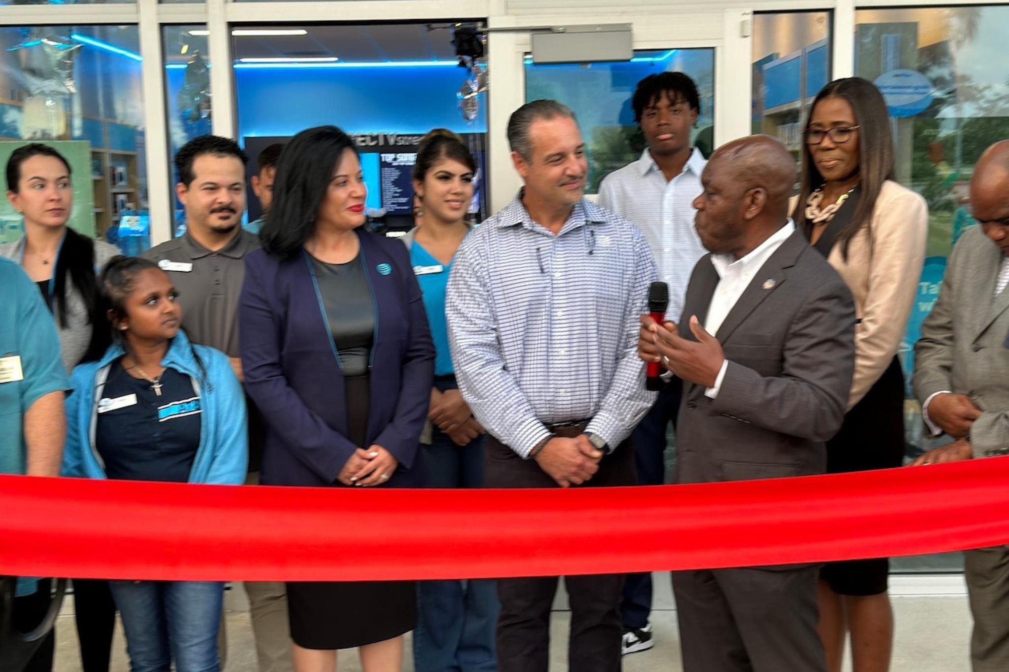 Business leaders and team members gathered at a retail store grand opening, with a speaker holding a microphone in front of a ceremonial ribbon.